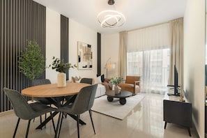 A modern living room with a round wooden dining table, grey upholstered chairs, a coffee table, a potted plant, a floor lamp, and a large window with sheer curtains. - Silkhaus New 1bdr Stay at Dubai Production City in Centrium Tower (Dubai)