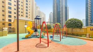 A playground with a yellow slide, red climbing structures, and a green climbing net, surrounded by high-rise buildings. - Silkhaus Iconic 2bdr Near Jumeirah Beach Resort (Dubai)