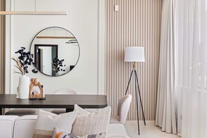 Ein modernes Wohnzimmer mit einem runden Spiegel, einem dunklen Holztisch, einem weißen Sofa mit gestreiften Kissen, einer Stehlampe und einer Vase mit getrockneten Blumen.