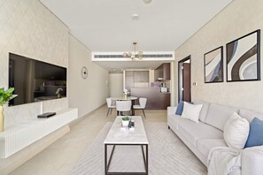 A modern living room with a white sofa, a coffee table, and a TV mounted on the wall. There is a dining area with a round table and chairs, and a kitchen in the background. - Silkhaus Deluxe 1BR Stay in Sky Gardens in Difc (Dubai)