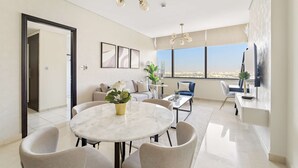 A modern living room with a round marble table, comfortable seating, a large window with a city view, and a flat-screen TV mounted on the wall.
