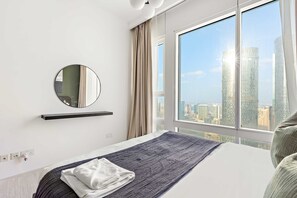 A modern hotel room with a large window, a bed with a grey textured bedspread, a round mirror, and a bedside table with a towel. - Silkhaus Chic 2BR Horizon Tower in Reem Island (Abu Dhabi)