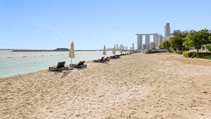 Una playa con tumbonas y sombrillas, cielo despejado y un horizonte urbano al fondo.