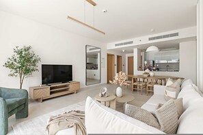 Ein modernes Wohnzimmer mit Flachbildfernseher, großem Spiegel, Essbereich mit Holztisch und -stühlen sowie gemütlicher Sitzecke mit Sofa und Kissen.