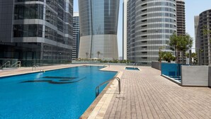 A swimming pool with a wooden deck, surrounded by modern high-rise buildings. - Silkhaus Brand New 1bdr Radiant Reem Island (Abu Dhabi)