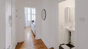 Ein Flur mit Holzboden, weißen Wänden, einem schwarz-weißen Schachfußboden im Badezimmer und einer weißen Tür links.