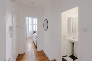 A hallway with a wooden floor, white walls, a black and white checkered floor in the bathroom, and a white door on the left. - Generous 2-bed Apartment Near Queen s Park (London)