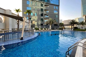 A swimming pool with a person in the water, surrounded by palm trees and modern buildings. - Stayshort - Stunning Studio Unit in Business Bay That Sleeps 2 (Dubai)