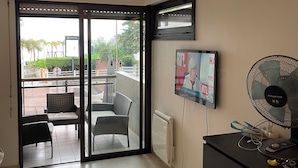 Ein Zimmer mit einer Schiebeglastür, die zu einem Außenbereich mit Korbmöbeln führt. An der Wand ist ein Flachbildfernseher angebracht und ein Standventilator steht auf einer Kommode.