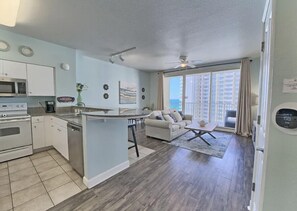 Eine moderne Küche mit einer zentralen Kochinsel, ein Essbereich mit einer Couch und einem Couchtisch sowie eine große Schiebeglastür, die auf einen Balkon mit Blick gelangt.