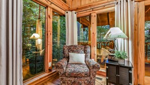 A wooden cabin interior with a patterned armchair, a small table with a lamp, and large windows offering a view of the forest. - Mountain Joy 1 Bedroom Cabin (Sevierville)