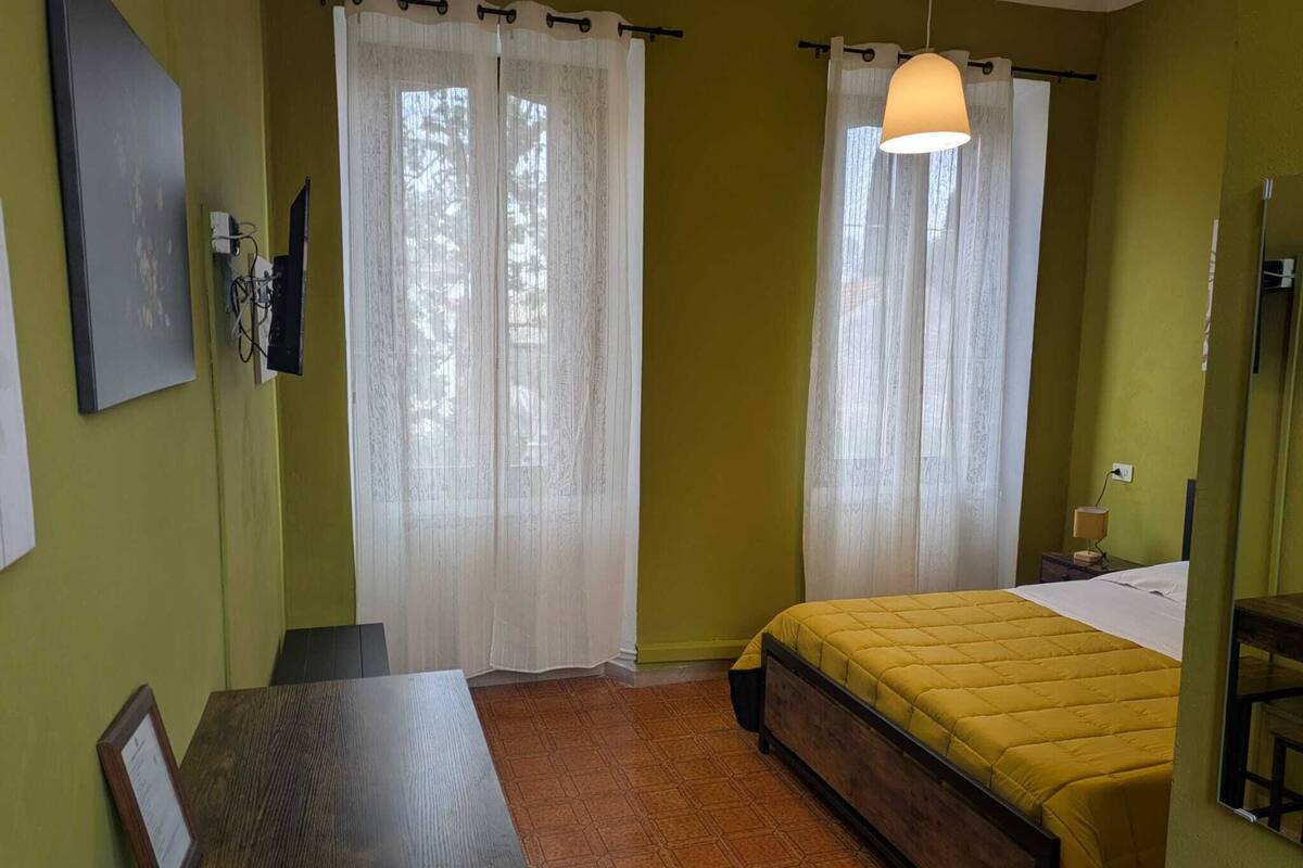Une pièce comprenant un lit, un bureau et une chaise. Les fenêtres sont dotées de rideaux et une lampe est suspendue au plafond.