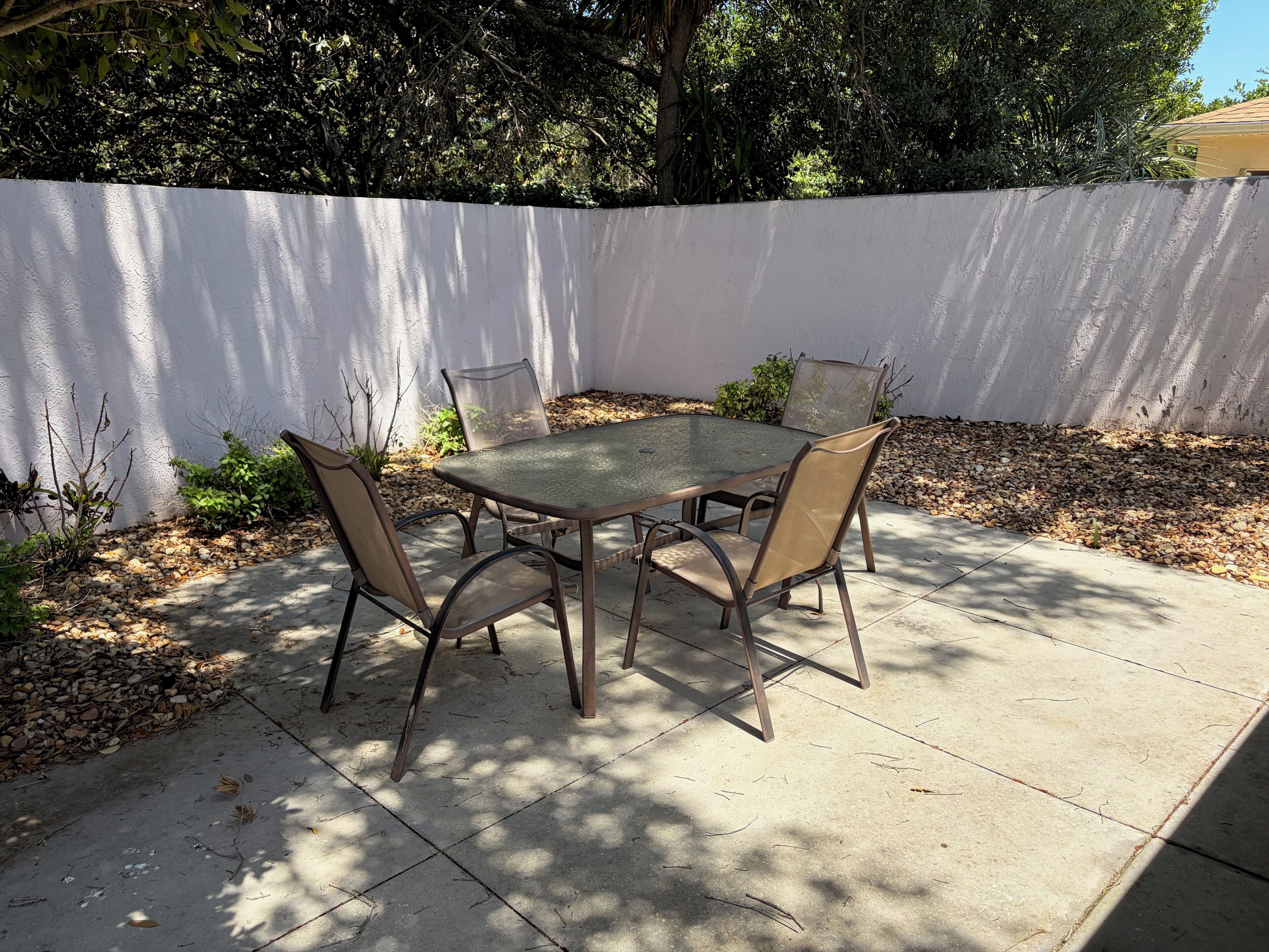 Un patio con una mesa de vidrio y cuatro sillas, rodeado por un muro blanco y vegetación.