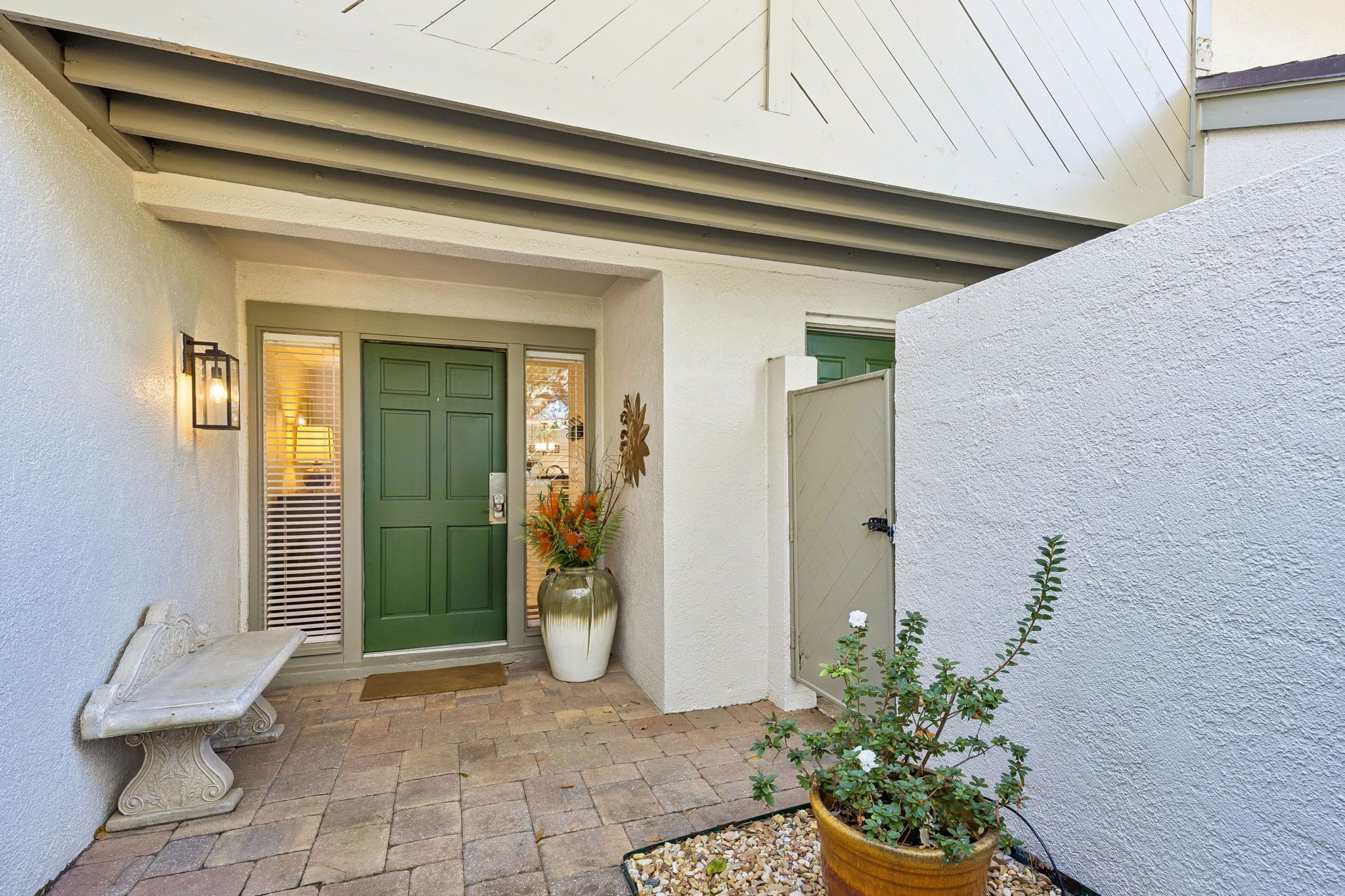 A courtyard with a green door, a bench, a large vase with flowers, and a potted plant.
