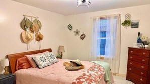 A bedroom with a wooden bed, a nightstand, a dresser, a window with curtains, and wall decorations including a hat rack and wall art.