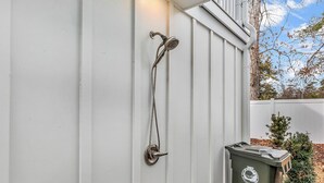 Eine Metall-Duschbrause an einer weißen Außenwand, ein weißer Zaun und ein grüner Mülleimer.