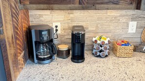Una cocina con armarios de madera, una cafetera, una máquina de café Keurig, un molinillo de café y un cesto con cápsulas de café.