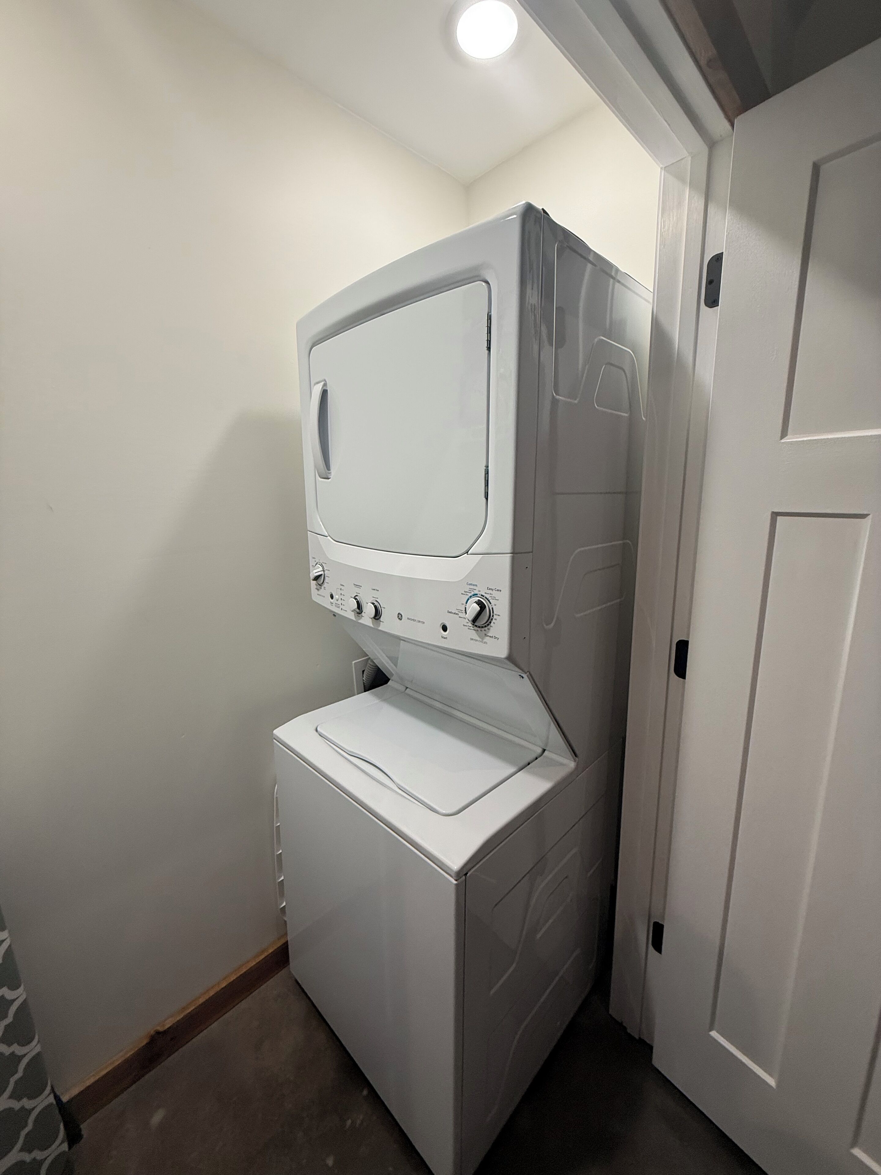 Een kleine, witte wasruimte met een voorlader wasmachine en droger, een bedieningspaneel met diverse instellingen, en een deur naar een andere kamer.