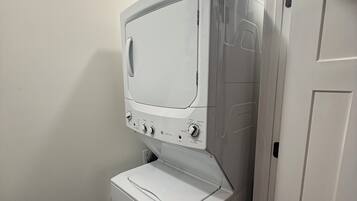 Een kleine, witte wasruimte met een voorlader wasmachine en droger, een bedieningspaneel met diverse instellingen, en een deur naar een andere kamer.