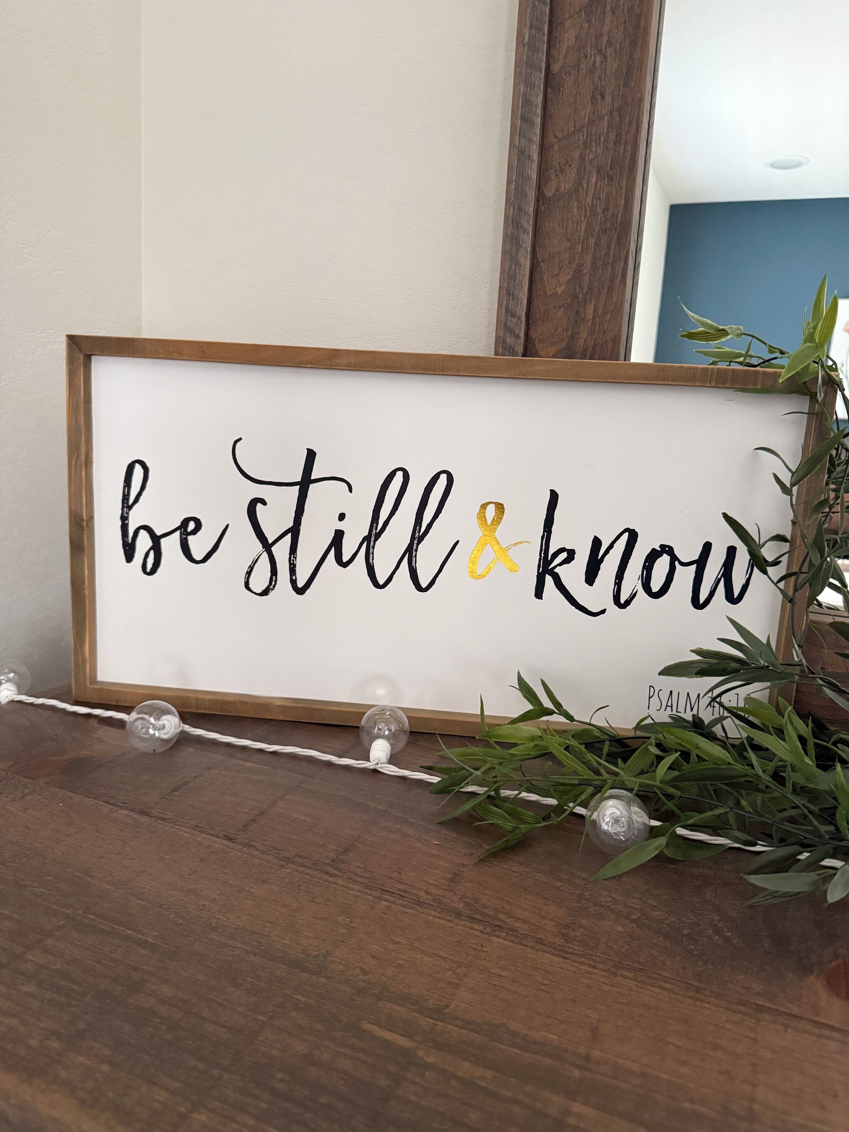Een lijst met een bord waarop staat 'be still & know' op een witte achtergrond, geplaatst op een houten oppervlak met een lichtslingering en begroeiing eromheen.