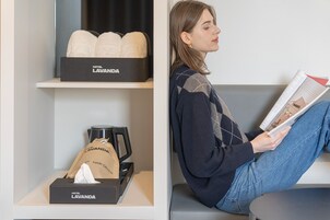 A hotel room with a shelf containing a box labeled 'Hotel Lavanda' and a hair dryer bag. A person is seated on a bench reading a magazine.