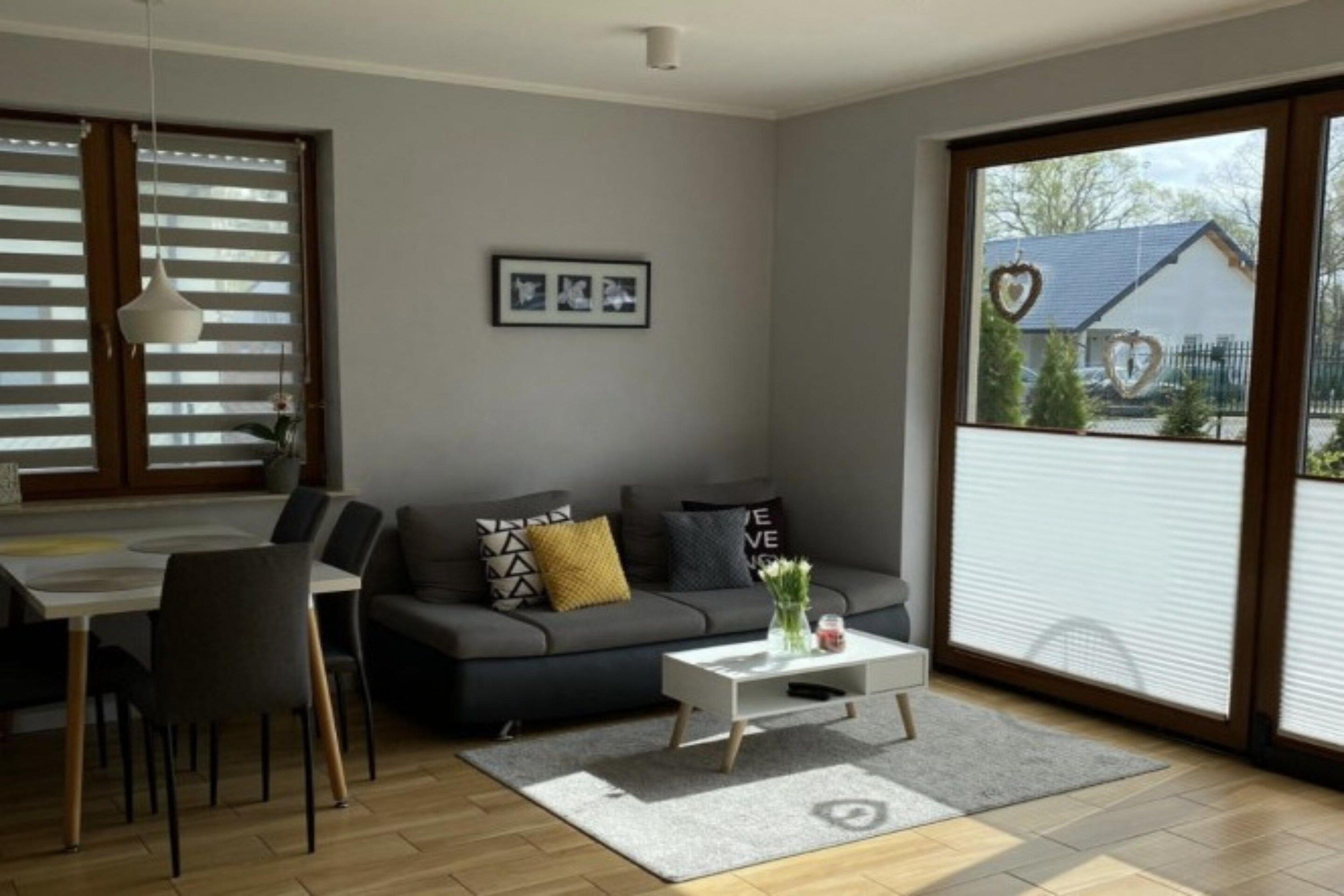 Un salon moderne avec un canapé gris, une table à manger avec des chaises, une table basse blanche et une grande fenêtre avec des stores.