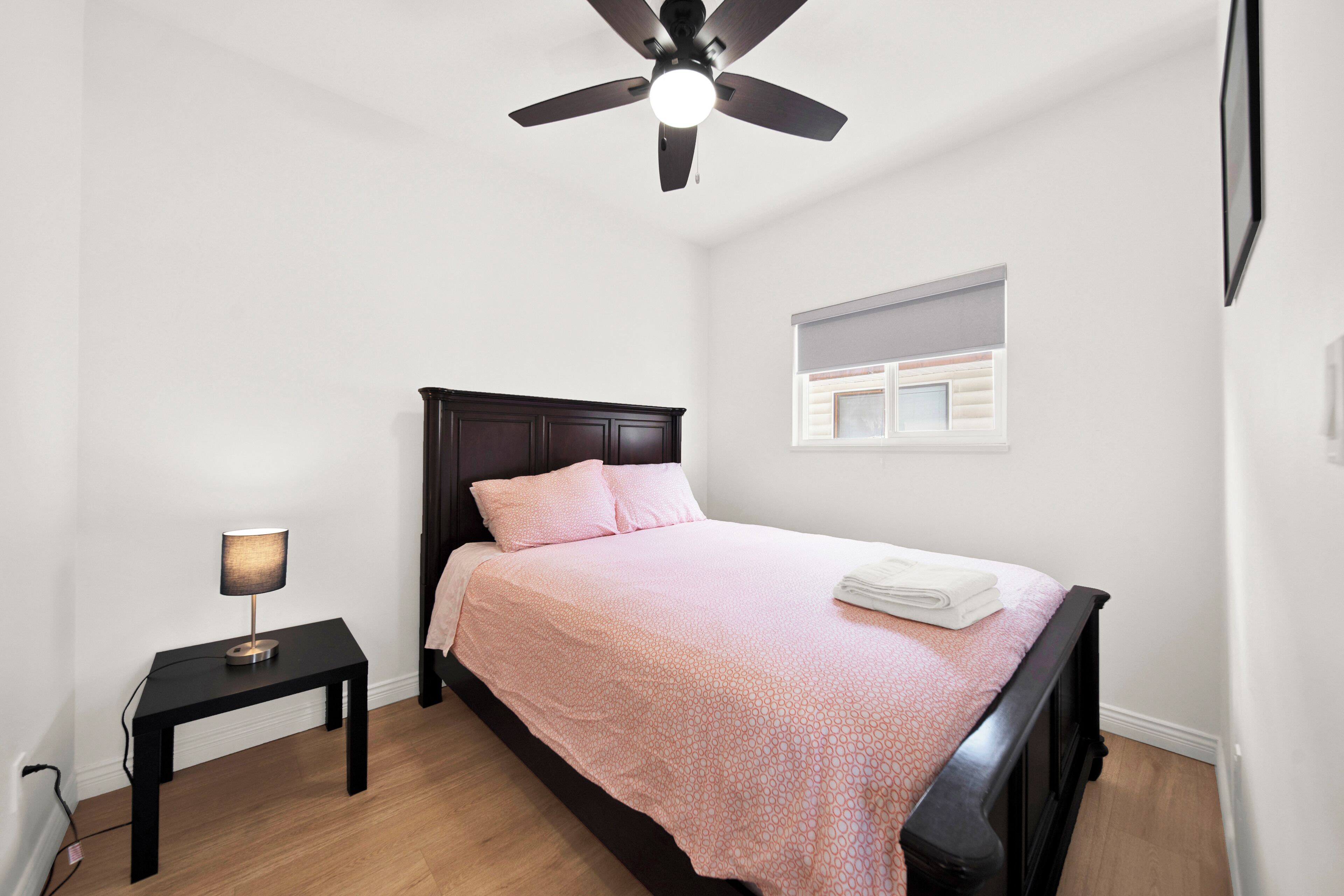 Une chambre à coucher comprenant un lit, une table de chevet, un ventilateur de plafond et une fenêtre avec des stores.