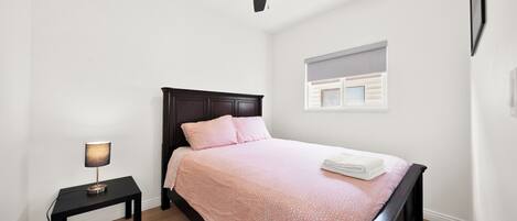 Une chambre à coucher comprenant un lit, une table de chevet, un ventilateur de plafond et une fenêtre avec des stores.