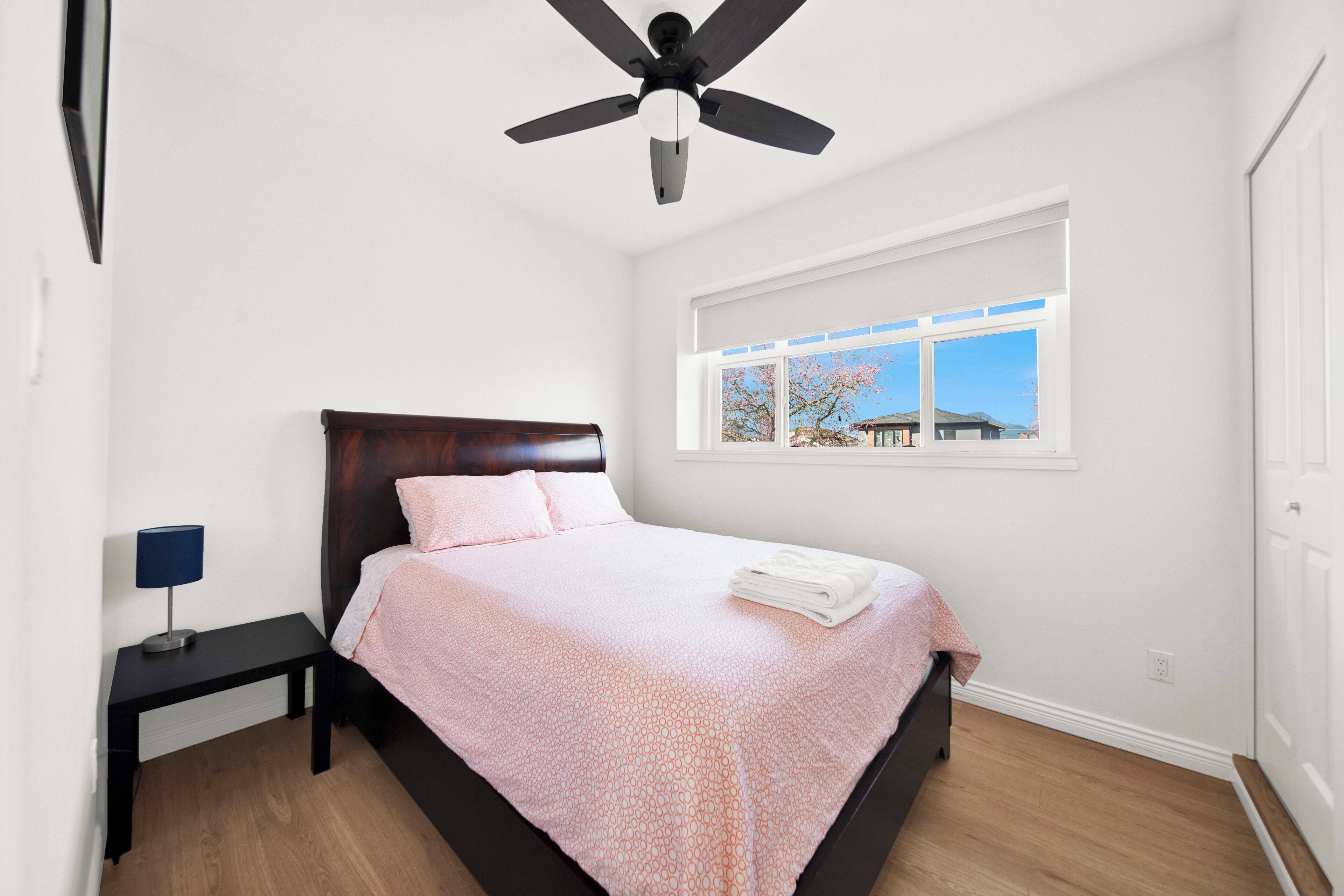 A bedroom with a bed, a nightstand, a ceiling fan, a window with a view, and a door.