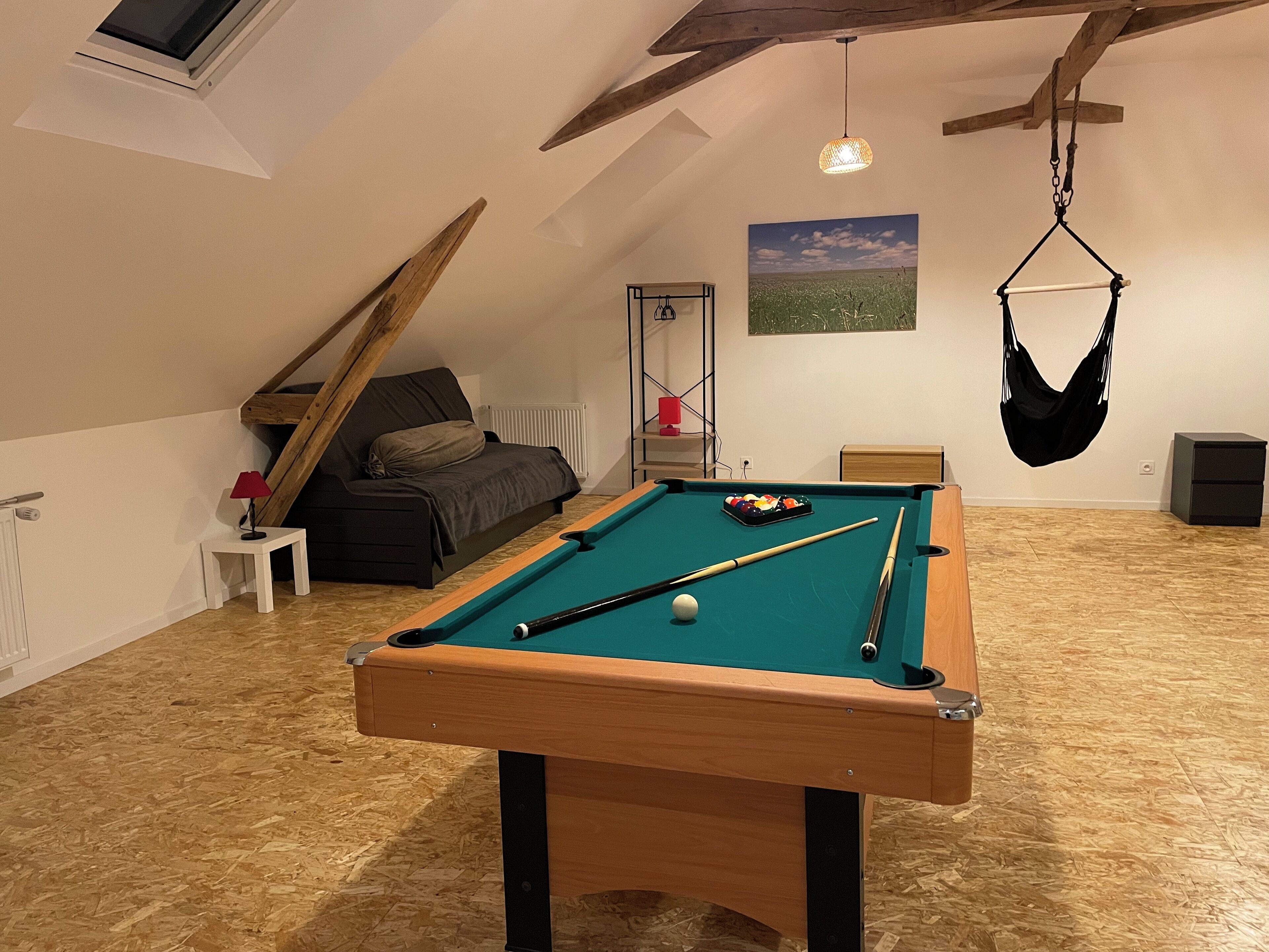 Une table de billard dans une pièce dont le plafond est en pente, avec un fauteuil suspendu et un tableau accroché au mur.