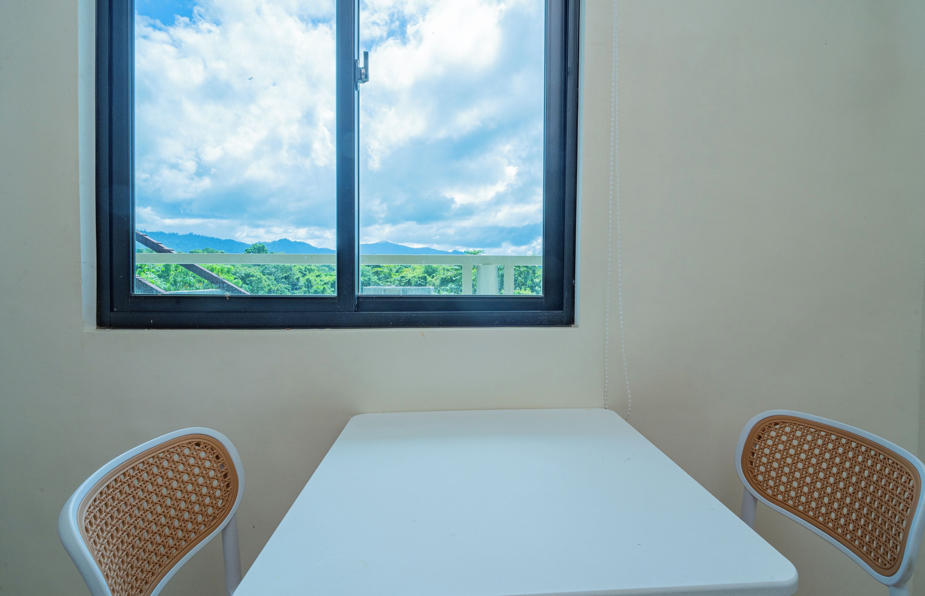 Quarto com mesa branca e duas cadeiras de vime, janela com vista para vegetação e montanhas.