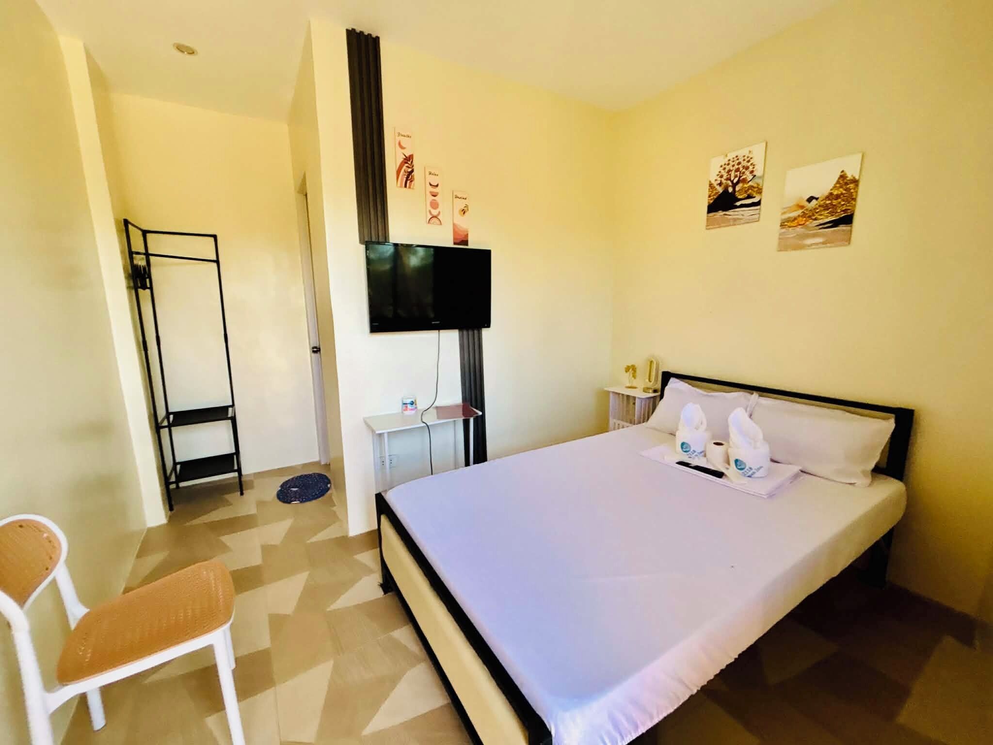 Une chambre d’hôtel avec un lit, une chaise, un téléviseur fixé au mur et une petite table sur laquelle se trouvent une bouteille et une tasse.