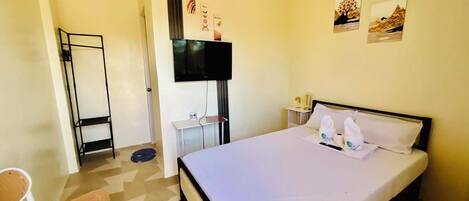 A hotel room with a bed, a chair, a TV mounted on the wall, and a small table with a bottle and a cup.