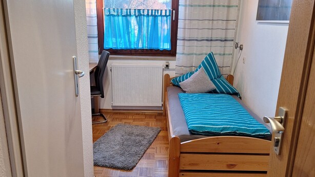 Un dormitorio pequeño y acogedor con una cama de madera, un radiador, una ventana con cortinas y vistas a árboles.