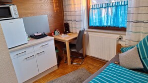 Habitación pequeña con kitchenette, escritorio, silla, cama y ventana con cortinas.