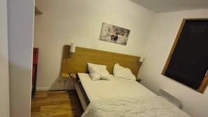 Een slaapkamer met een houten vloer, een bed met wit beddengoed, een hoofdbord, een aan de muur bevestigd schilderij en een raam met een jaloezie.
