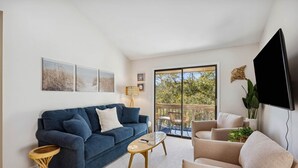 Een woonkamer met een blauwe bank, een beige fauteuil en een salontafel. Er is een televisie aan de muur bevestigd en een raam met uitzicht op groen.