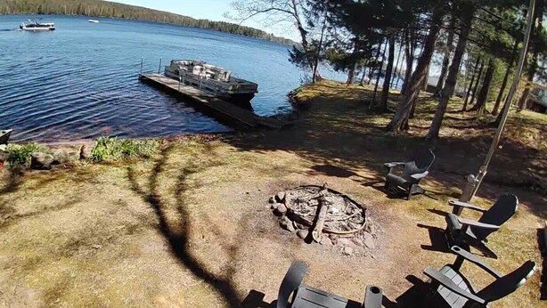 Ein Campingplatz am Seeufer mit Feuerstelle, Stühlen und einer Anlegestelle mit einem Boot.