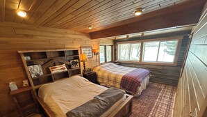 Ein gemütliches Schlafzimmer im Chalet-Stil mit einem großen Bett, einem kleineren Bett und einem Nachttisch mit Lampe. Ein Fenster bietet Blick auf Bäume, und ein Schild informiert über nordamerikanische Fische.