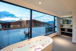 Une salle de bain moderne dotée d’une grande baignoire, d’une cabine de douche en verre et offrant une vue sur un bâtiment traditionnel grâce à de grandes fenêtres.