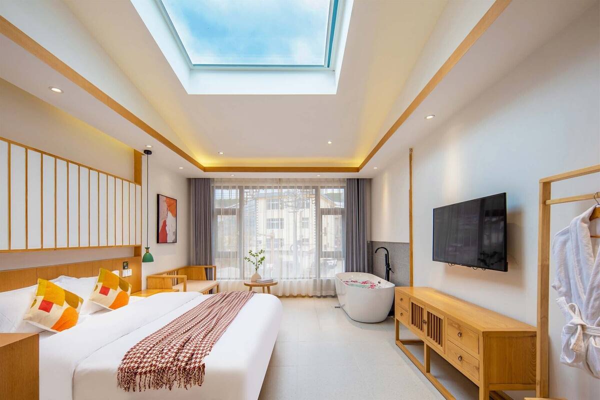 A modern hotel room with a skylight, a large bed, a bathtub, a TV, and a wooden vanity.