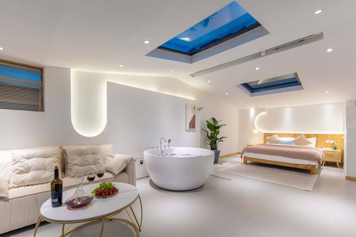 A modern bedroom with a skylight, a large bathtub, a sofa, and a bedside table with a bottle and grapes.