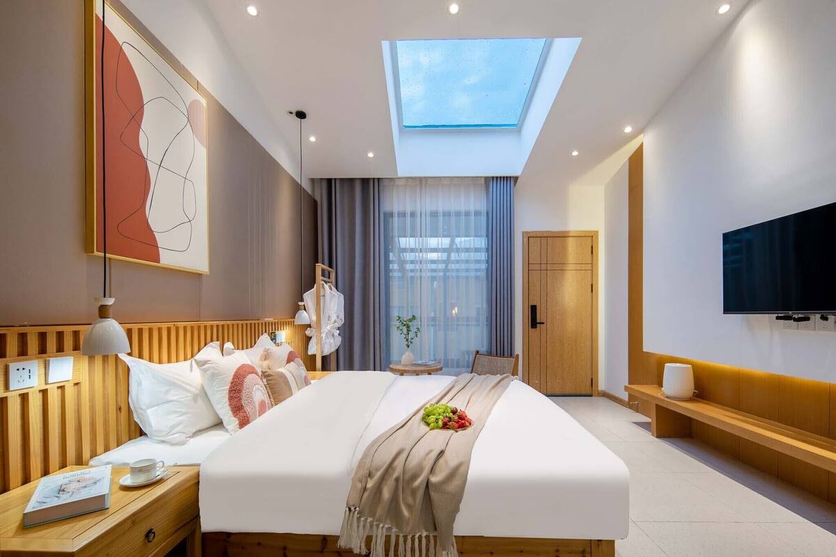 A modern hotel room with a skylight, a large bed, a bedside table with a book, a TV, and a wooden headboard.