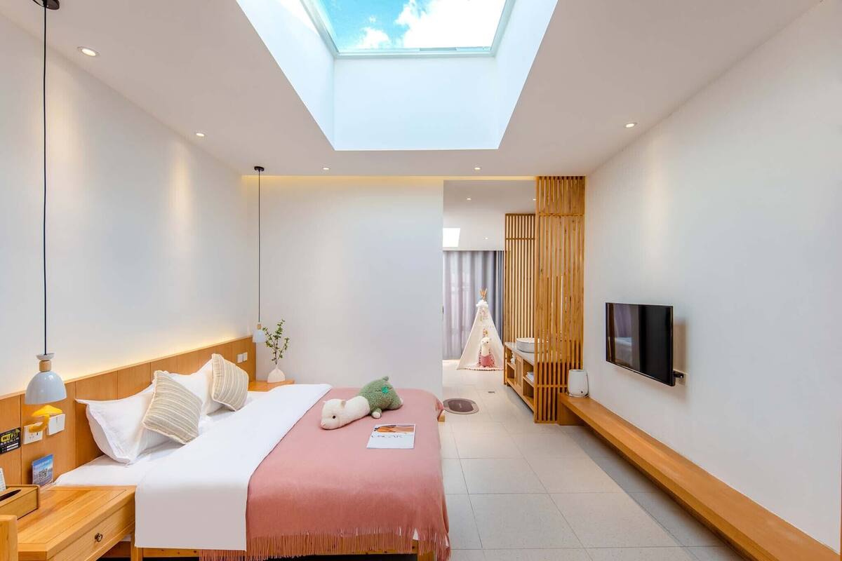 A modern hotel room with a skylight, a bed with pillows, a bedside table, a TV mounted on the wall, and a small wooden shelf.