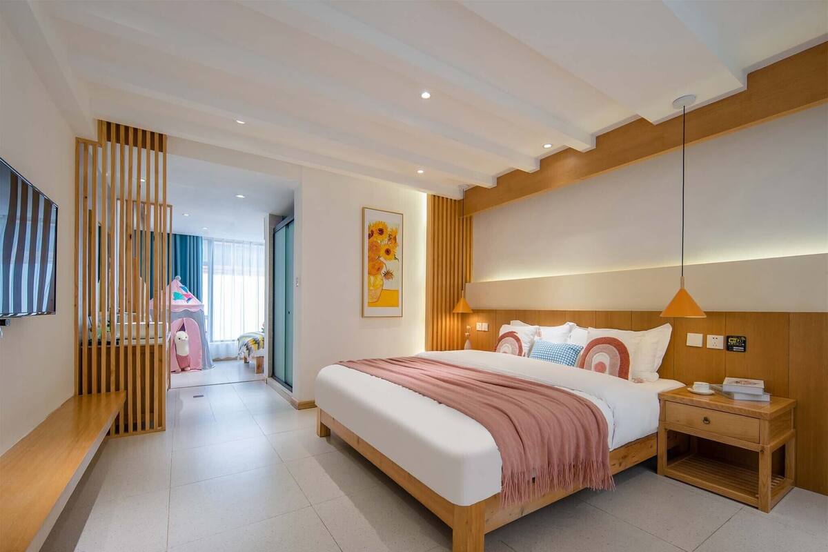 A modern hotel room with a large bed, wooden headboard, and a nightstand. There is a wooden partition with a window, a wall-mounted TV, and a framed artwork above the bed.