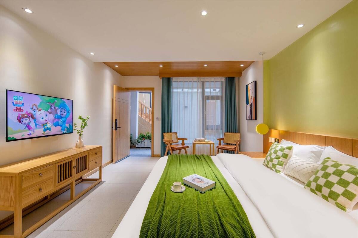 A hotel room with a bed, a TV, a wooden console, a green blanket, and a window with curtains.