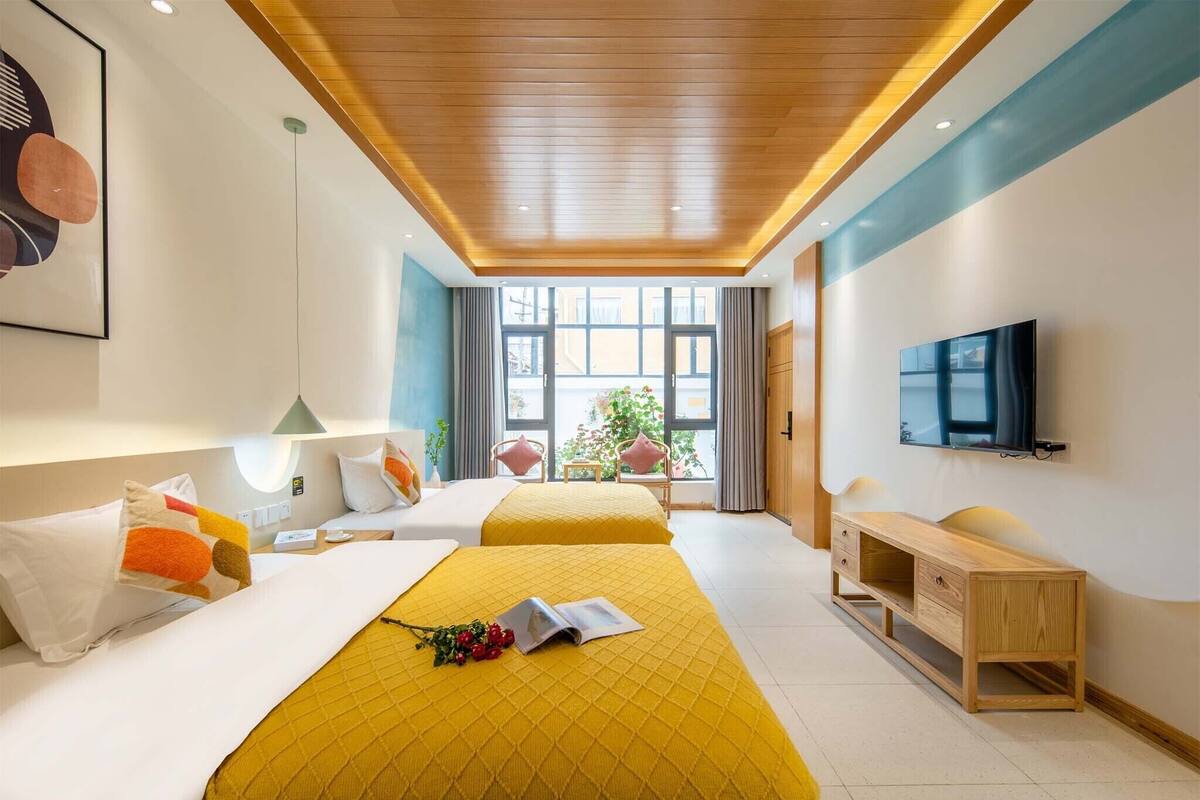 A hotel room with two beds, a TV, a wooden desk, and a window with a view of greenery.