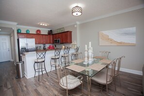 A modern kitchen with wooden cabinets, stainless steel appliances, and a glass dining table with metal chairs. - Gulf-Front 2BR • Sleeps 6 • Ocean Villa 702 • Master w/Amazing Views (Panama City Beach)