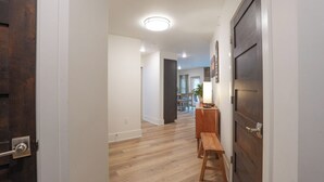 Um corredor com piso de madeira, uma porta de madeira escura à direita e um banco com mesa e abajur.