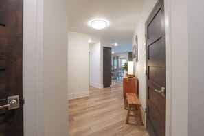 A hallway with wooden flooring, a dark wooden door on the right, and a bench with a table and lamp. - Blue River Flats Basecamp Condo: Walk to Blue Bird Market & Shops (Silverthorne)